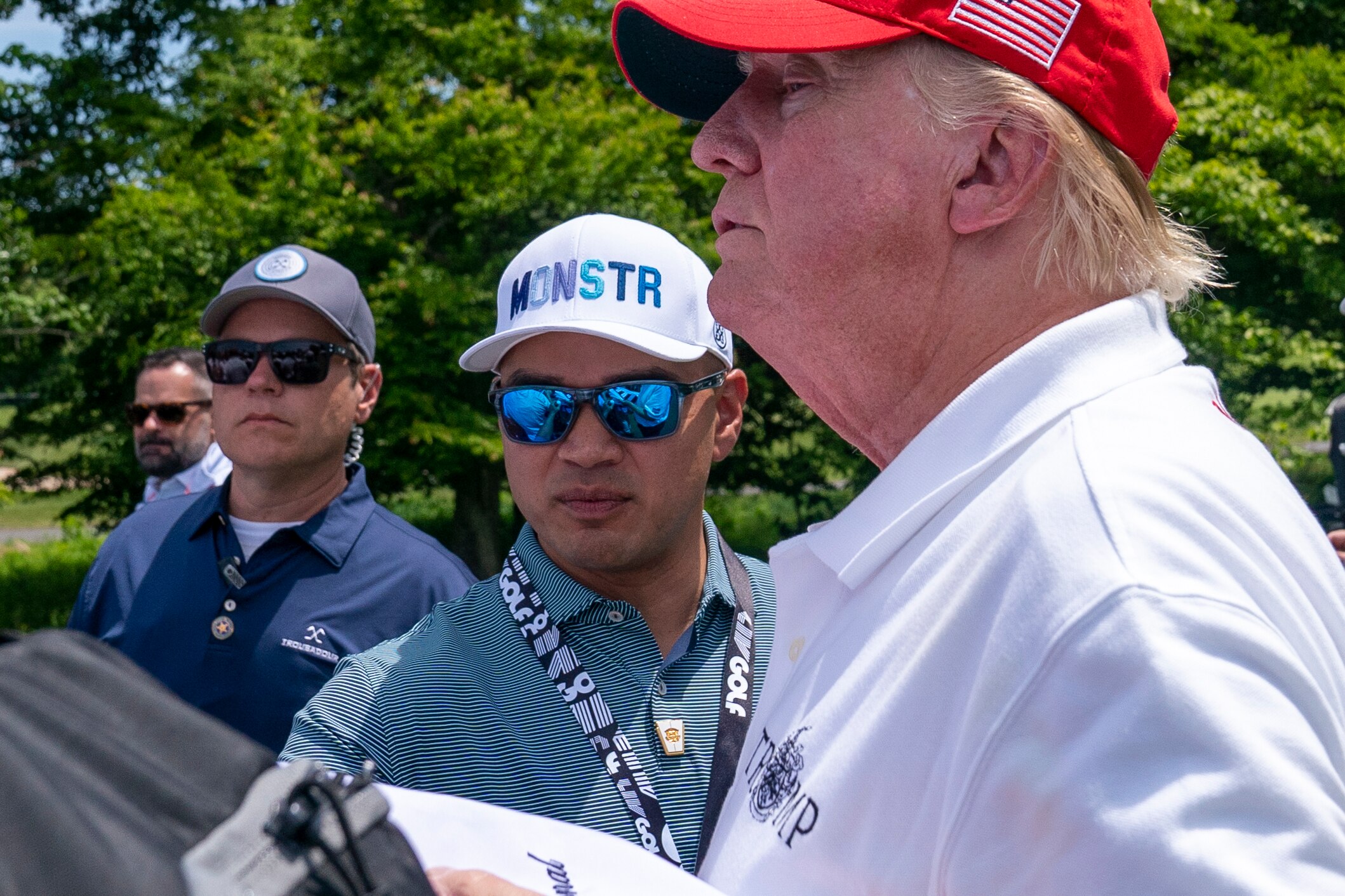 A man wearing sunglasses, a lanyard and a cap stands beside Donald Trump at a golf course.
