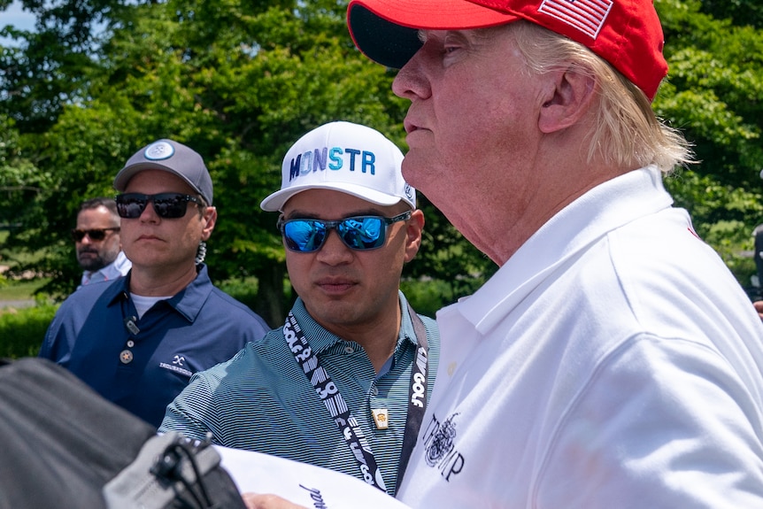 A man wearing sunglasses, a lanyard and a cap stands beside Donald Trump at a golf course.