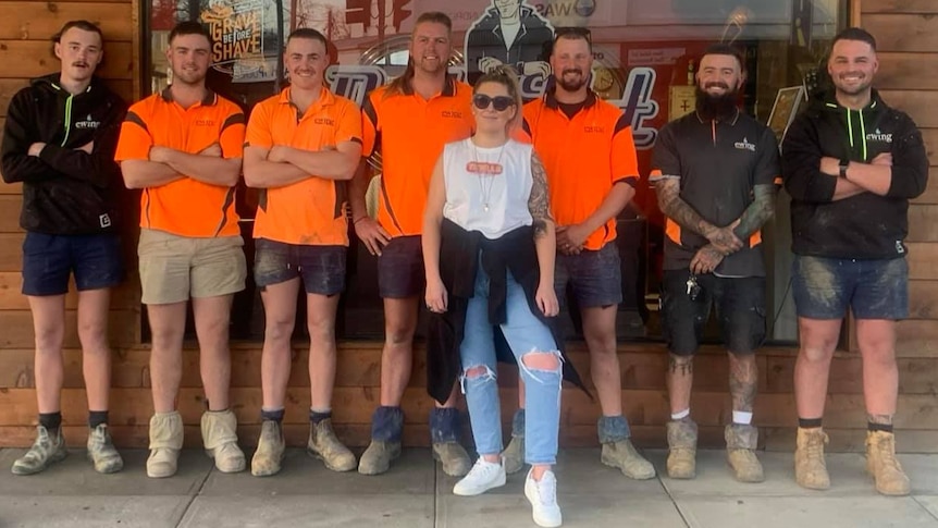 A group of men, wearing orange tradie shirts, and one woman stand in a line
