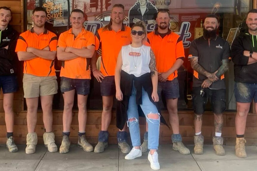 A group of men, wearing orange tradie shirts, and one woman stand in a line