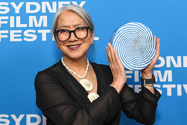 A woman in a sheer black top, a large necklace & glasses holds an award in front of a photowall reading "Sydney Film Festival",