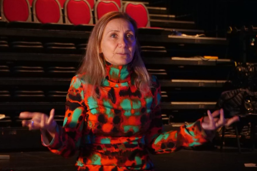 Deborah Leiser-Moore holds her hands out as she stands in front of the audience seats inside the theatre.