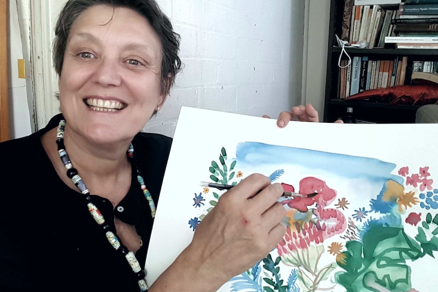 A smiling woman with short hair holds up a vibrant watercolour painting she has created.