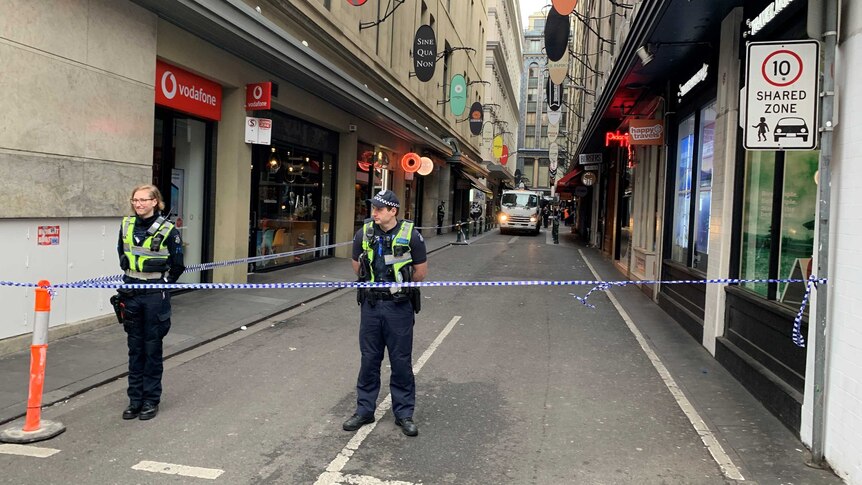 Police stand behind police tape on a laneway, with a rubbish truck behind them.