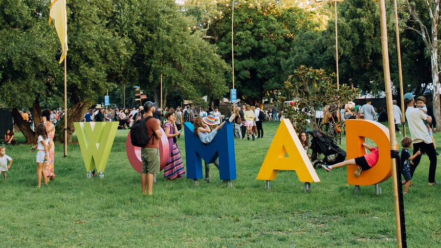 Crowds of people gather in a park. Children play in large letters that spell WOMAD