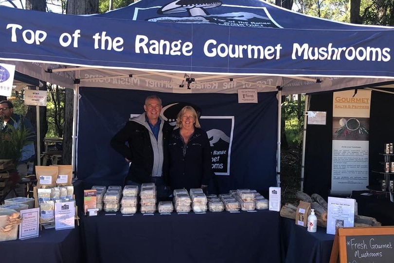 Man and Female stand under a gazebo at a market stall selling mushrooms