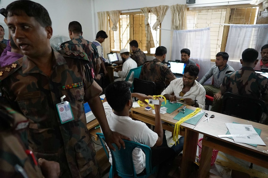 Rohingya men sit around a room with officials and laptop computers.