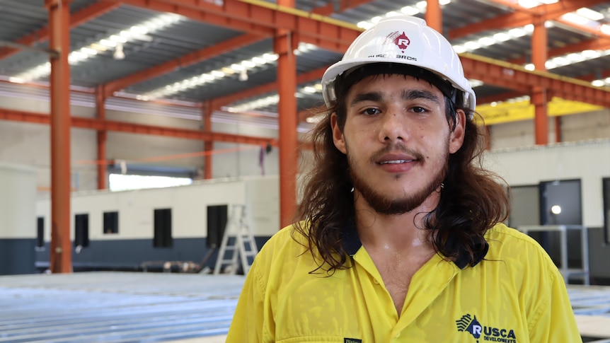 A man in his 20s, wearing a high vis shirt and hard hat, standing in a warehouse and looking at the camera.