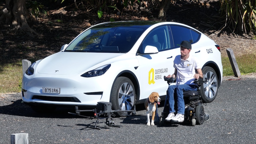 a man in a wheelchair sits beside a Tesla