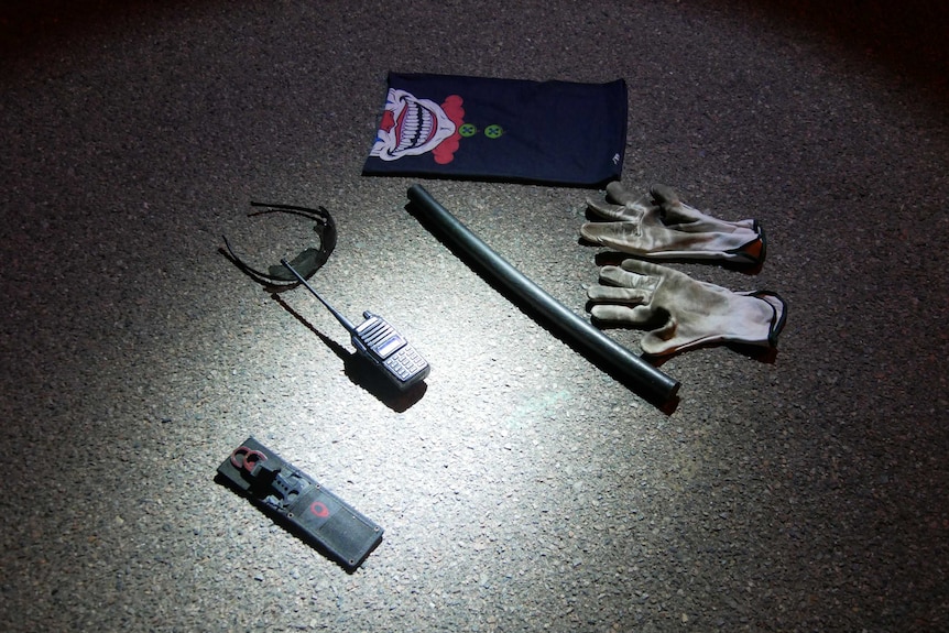 A clown bandana, gloves, baton, sunglasses and walkie-talkie lying on asphalt.