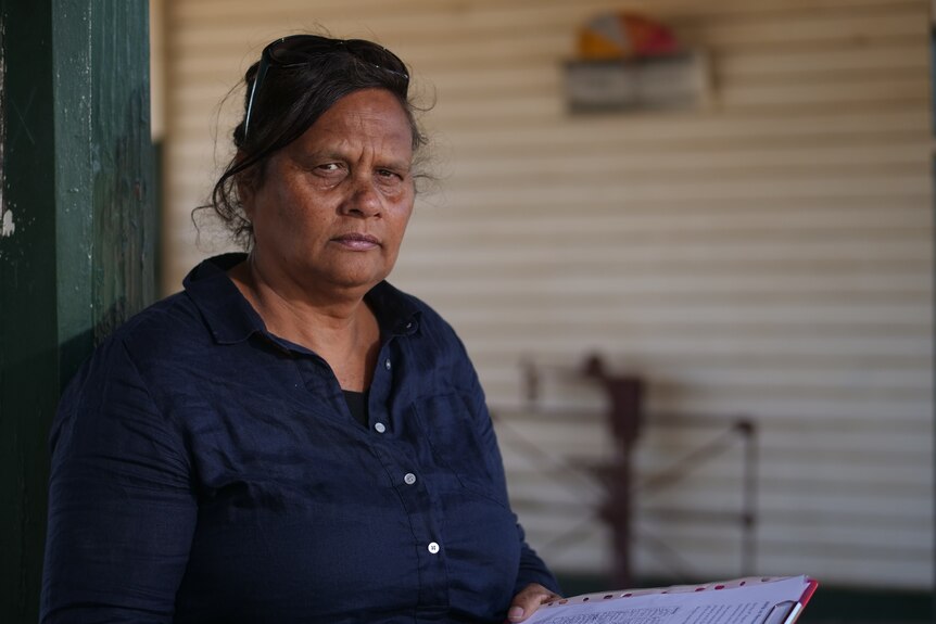 An Aboriginal woman with a clipboard, wears a navy blue shirt, sunglasses on her head, looks seriously at camera.