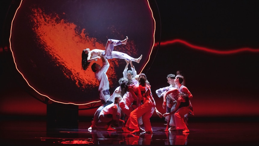 A group of dancers in white on a stage. One dancer is lifted into the air. A giant sun-shaped image is projected behind them.