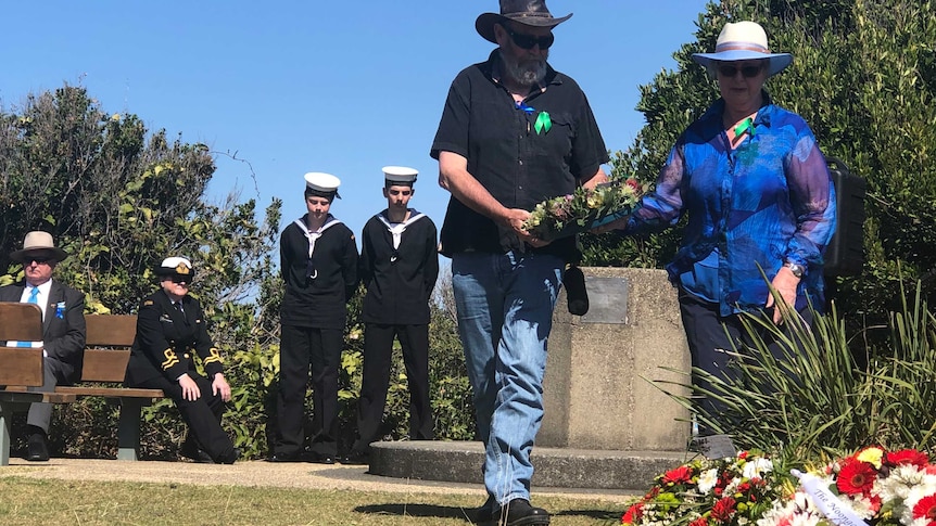 Two people place a wreath on the ground with boys dressed as sailors in the background