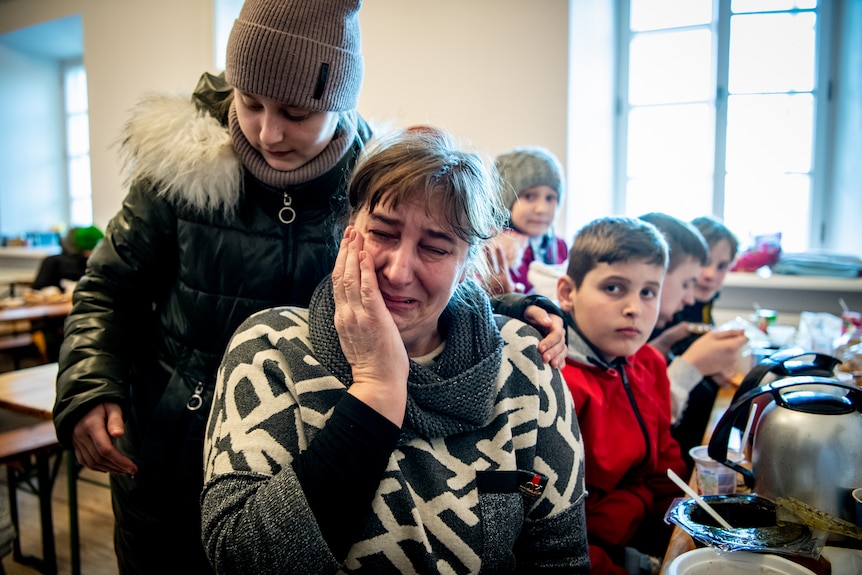 A woman crying, with her hand on her face, as a boy in a beanie comforts her