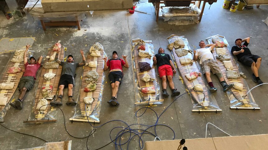 Six men lay on the ground in a workshop next to six wooden surfboards piled with sandbags.