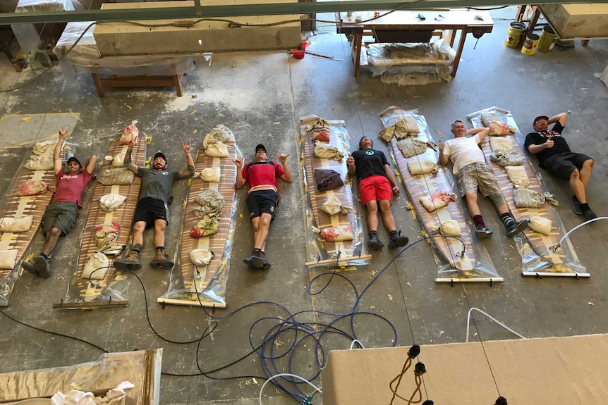 Six men lay on the ground in a workshop next to six wooden surfboards piled with sandbags.