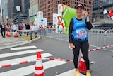 A women in running clothes standing an intersection in a big city, smiling and holding a medal.
