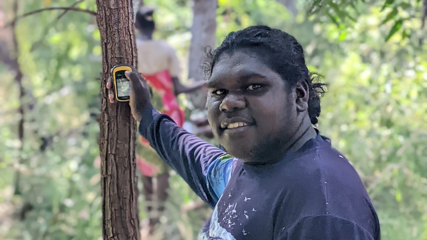 A young aboriginal man in a dark blue long sleeve shirt holding a gps up to a tree outside. Smiling at camera.