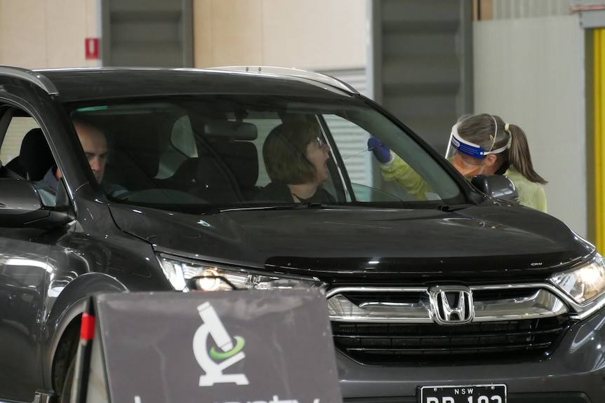 A man and woman sitting in a car, the woman is being swabbed by a nurse 