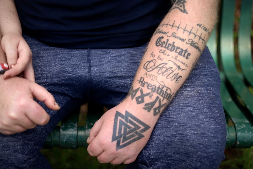 A tattoo on a man's arm reads 'celebrate this chance to be alive and breathing', with his son's name and heartbeat above it.