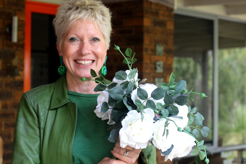 A woman wearing a green jumper stands holding a bunch of white flowers to one side.