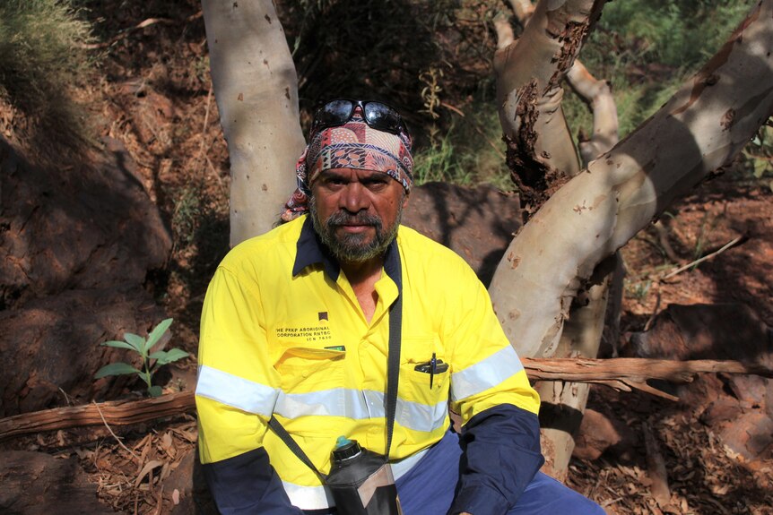 An Indigenous man in a bandana and a high-vis shirt sitting in bushland, looking determined.