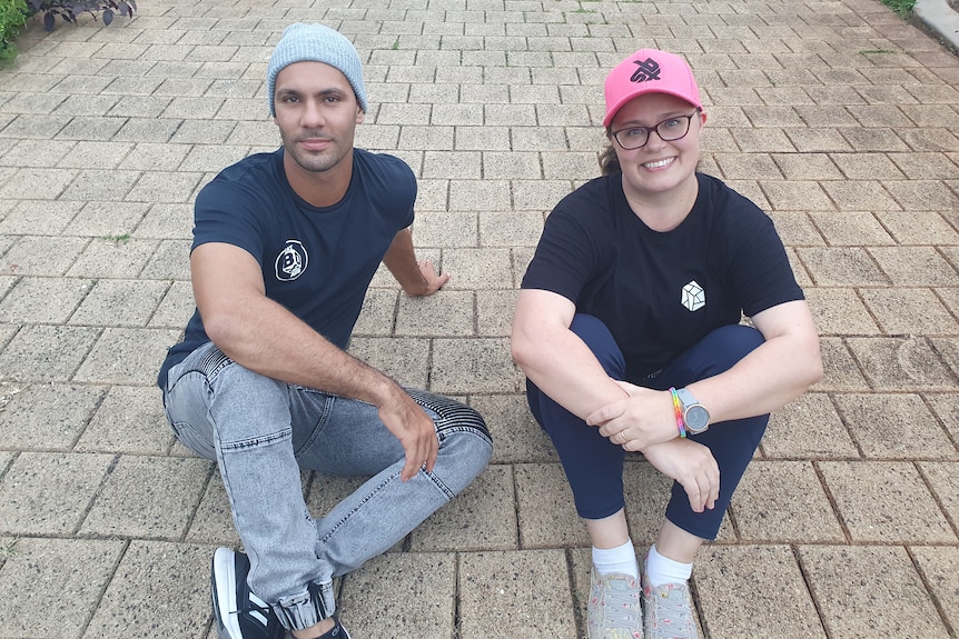 A man in a beanie and a woman in brightly coloured cap sit side by side on a driveway.