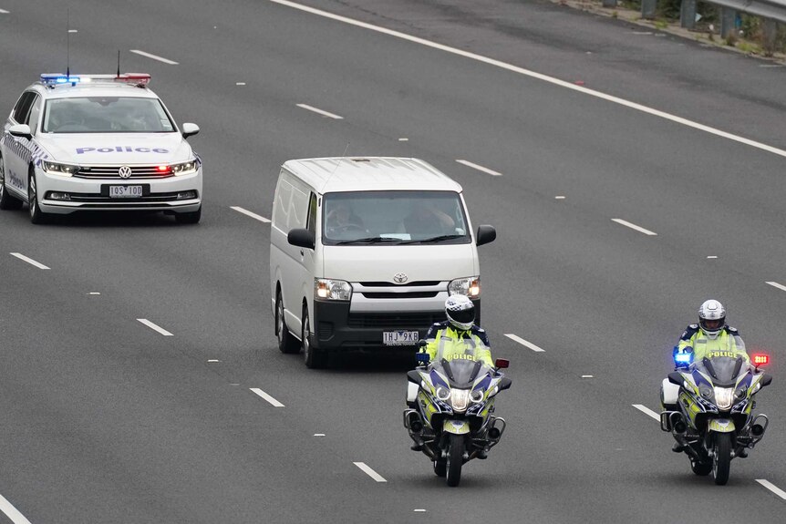 Two police motorbikes and a police car escort a white van on an empty road.