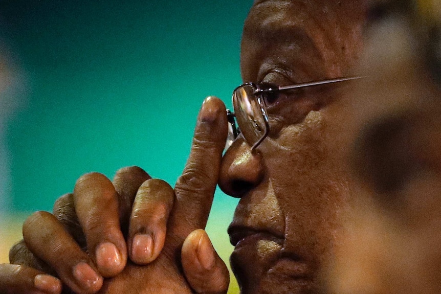 Profile view of Jacob Zuma, sitting expressionless with hands clasped in front of him.