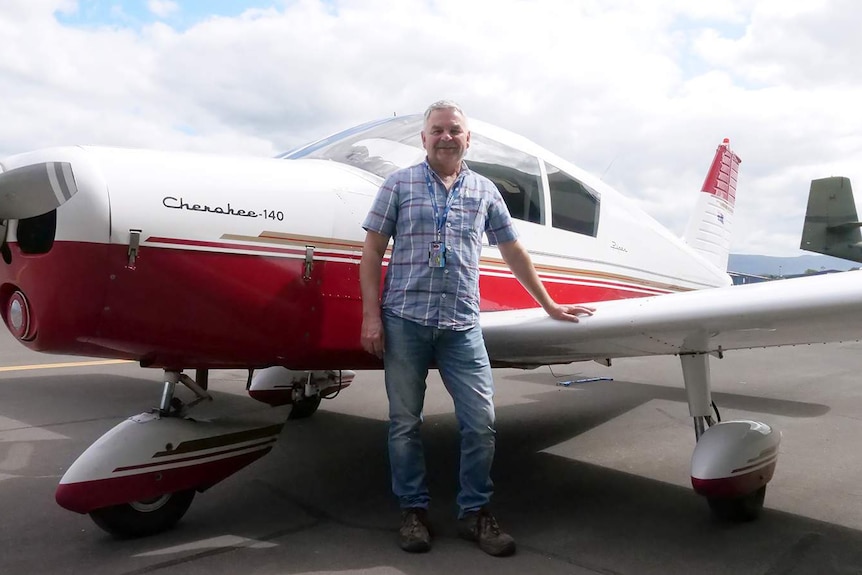 Mark Keech stands in front of a red and white Cherokee 140 aeroplane with his hand resting on a wing.