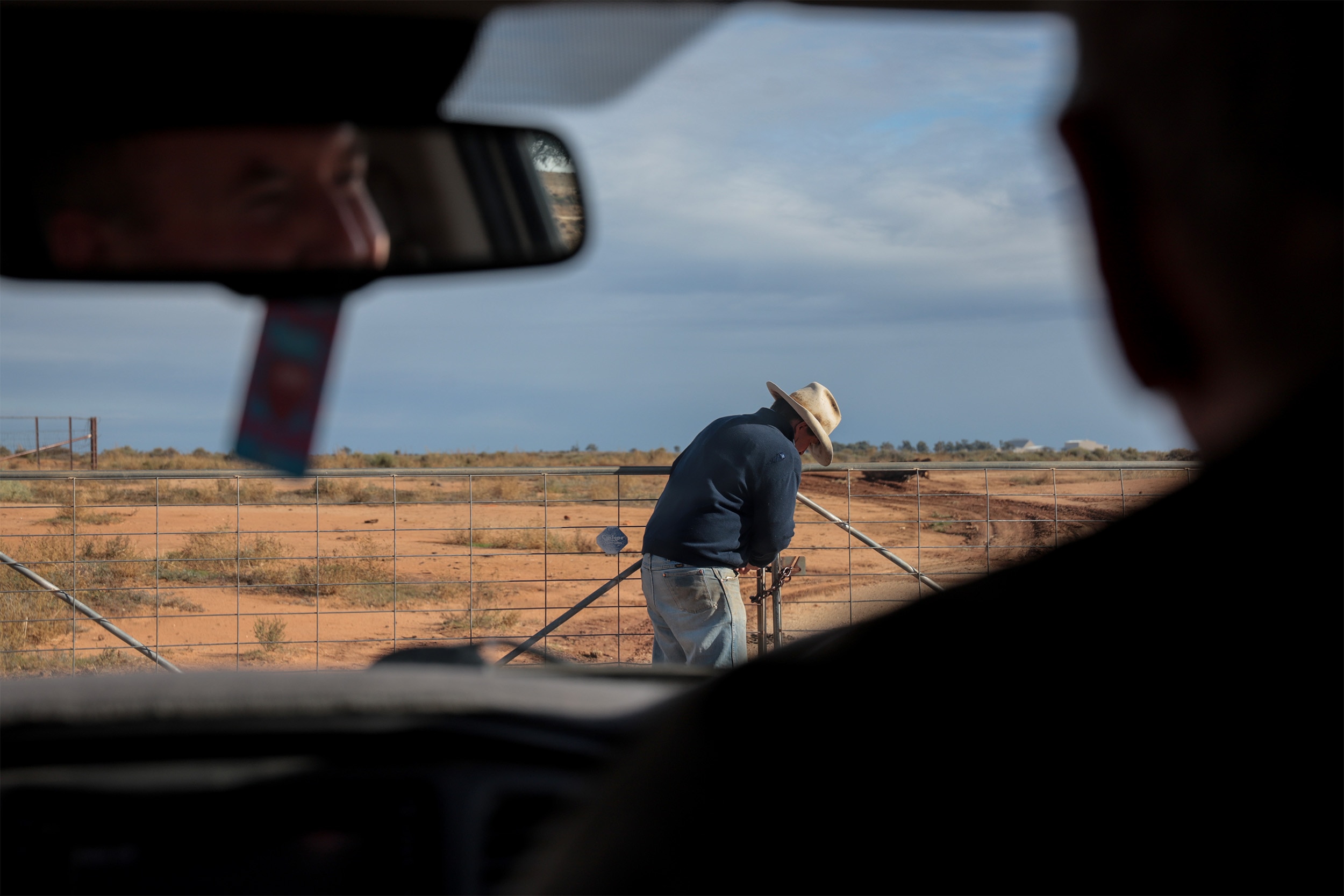Man wearing weathered hat opens a farming fence on a country property while silohuetted man in a car watches on