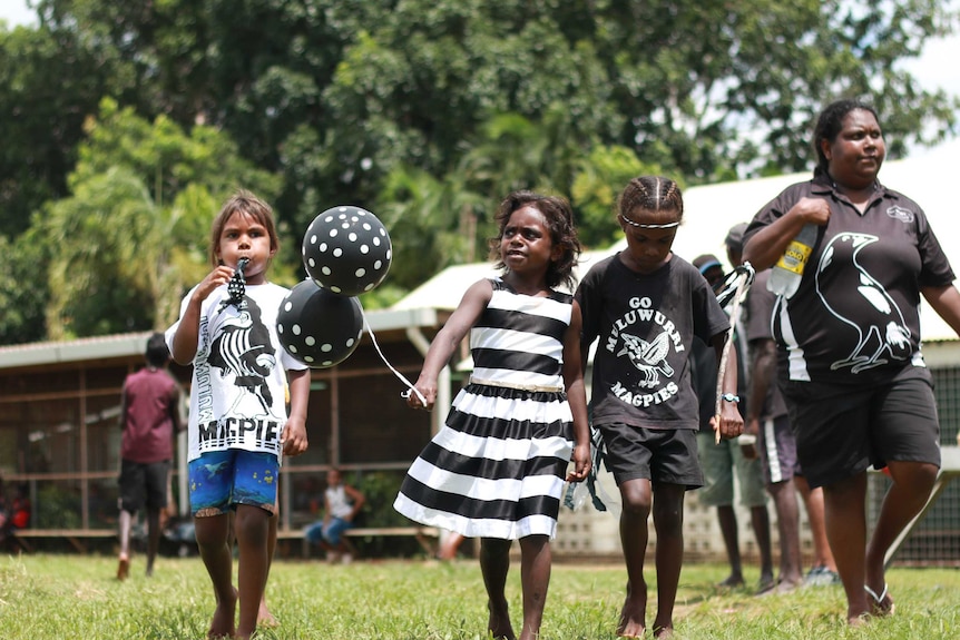 Group of girls wearing Muluwurri Magpies shirts and with black and white balloons walk in a line at the final.