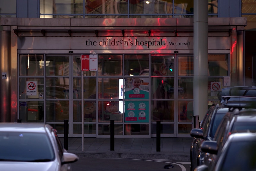 An entrance to a hospital. Cars are parked outside it. Lights reflect off the building's metallic surfaces.