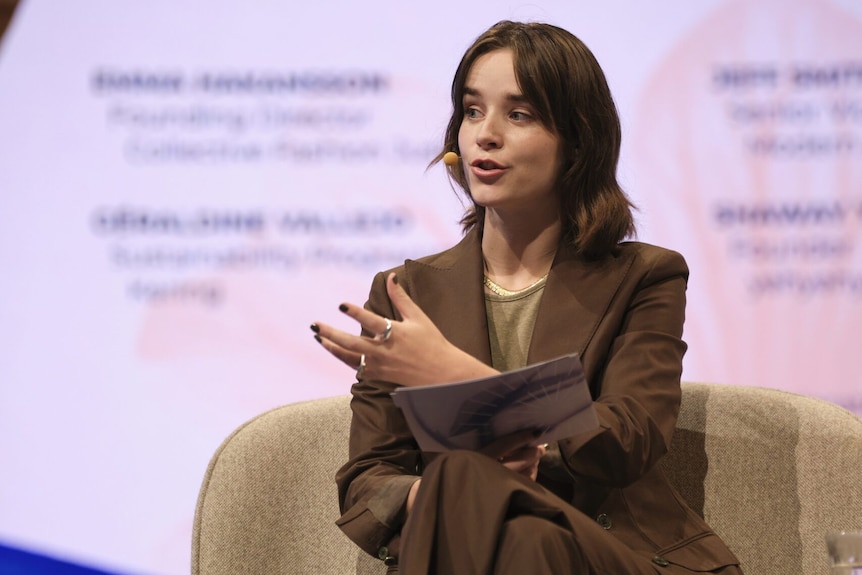 Emma, a fair-skinned young brunette woman, wears a blazer and gesticulates in a chair at a conference. 