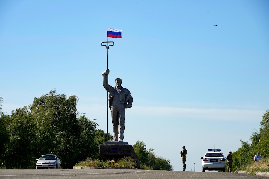 A Russian soldier stands in the road at the entrance of Mariupol with a Soviet style symbolic monument of a metallurgist. 