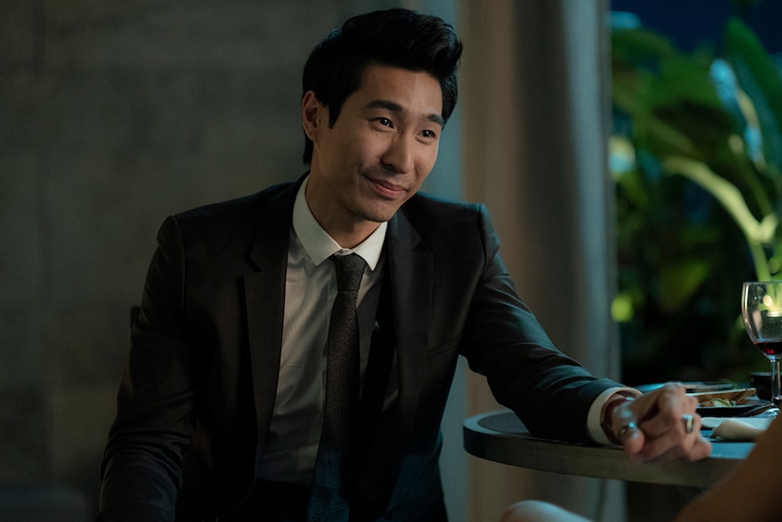 Chris Pang sits with arm resting on dining table with plates and wine glass and wears black suit indoors in evening.