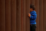 A small child holds on to large rusty metal bars that form a wall along the US/Mexico border