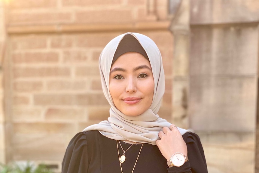 A woman with her head wrapped in a cream scarf, with a black top on, pensive expression in front of a stone building