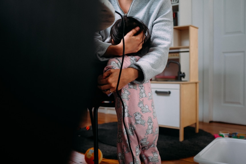 Partly obscured by a dark table a person visible from shoulders down holds a small child.