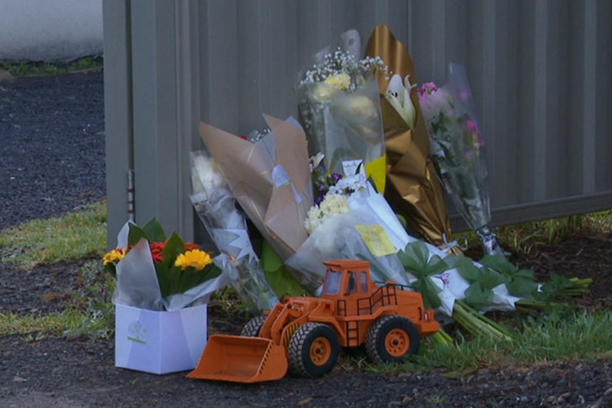 Bunches of flowers and a toy excavator are left in front of a metal fence.