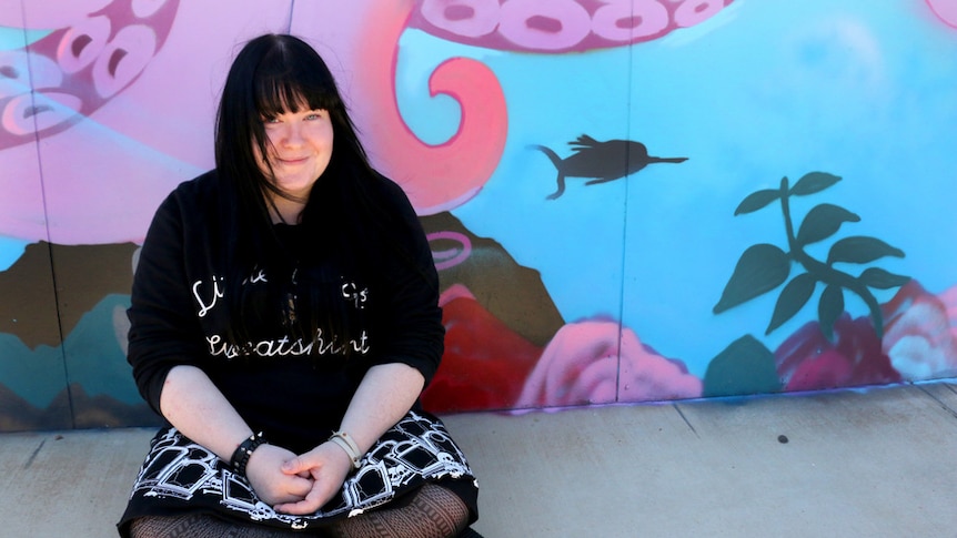 A girl with long black hair, dressed in a black skirt covered with skulls, sits in front of a wall.