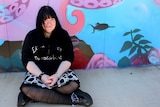 A girl with long black hair, dressed in a black skirt covered with skulls, sits in front of a wall.