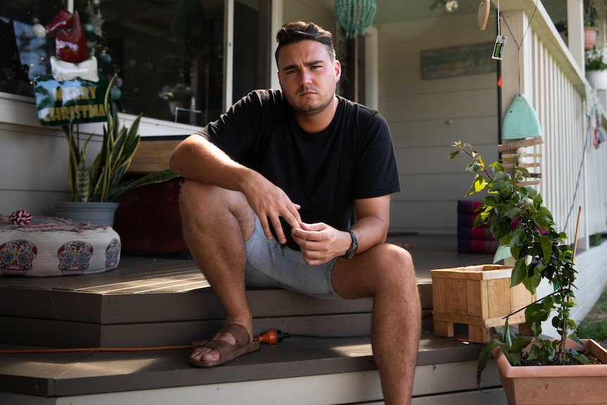 A man sits on the steps of a front porch, wearing a black tshirt and sandals. He looks dejected. 