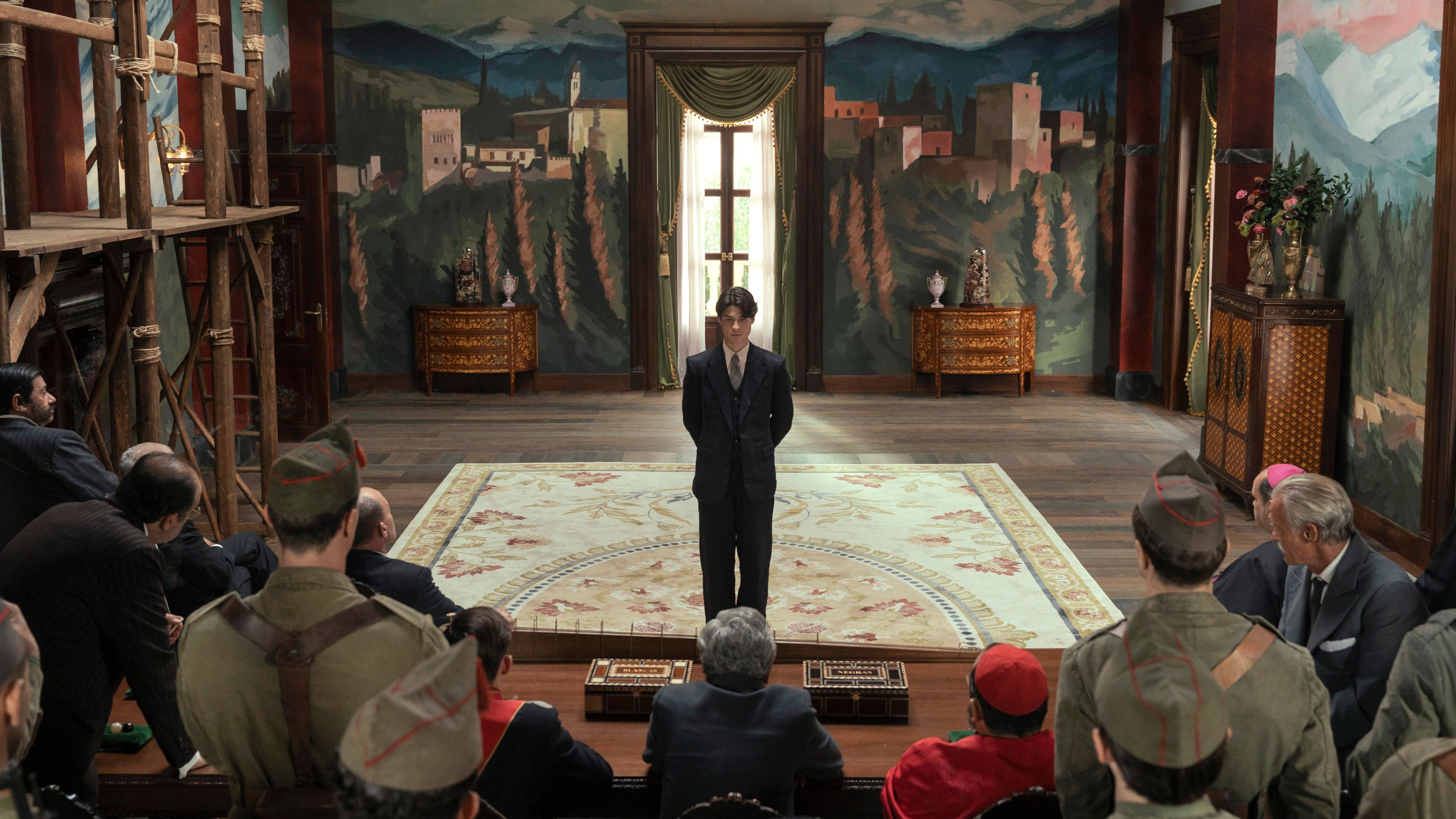 A young man in a black suit stands in front of a group of older men in a baroque building