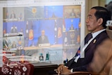 Joko Widodo sitting back in a chair with a projection of ASEAN virtual meeting