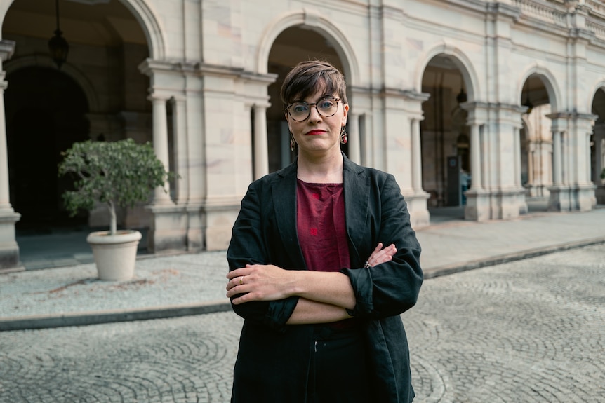 A woman with glasses stands with arms folded. 