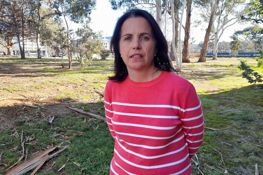 Mandy looks serious, standing outside near trees, wearing a pink and white jumper.