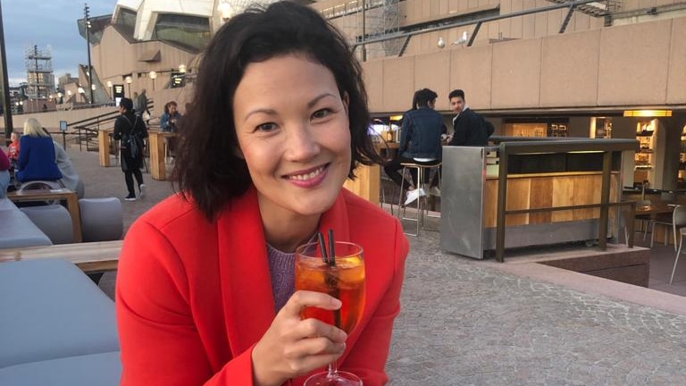 Comedian and writer Lizzy Hoo sitting outside Sydney Opera House with a cocktail.