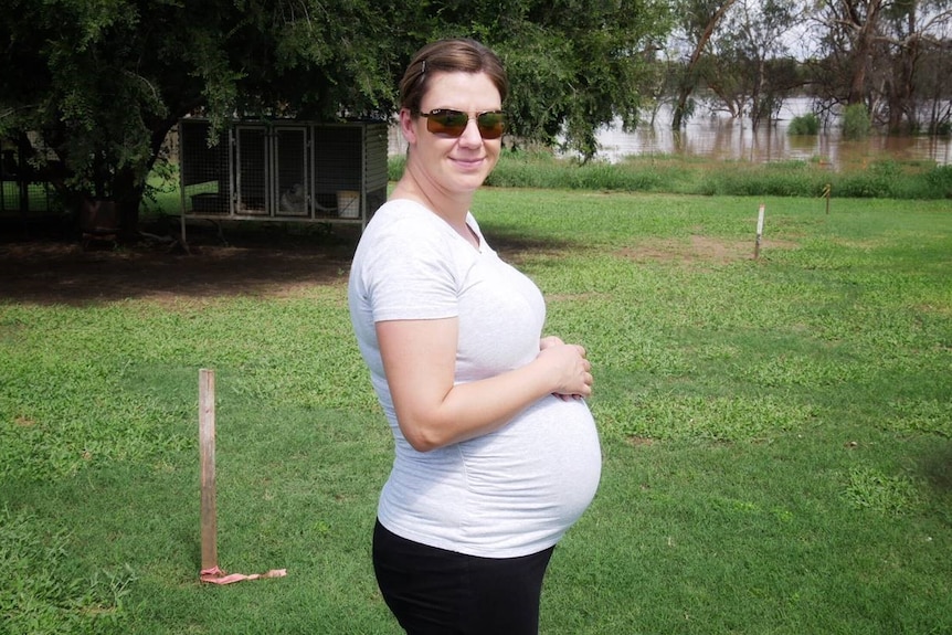 A pregnant woman wearing a shirt and sunglasses rests her hands on her pregnant belly with a flood in the background