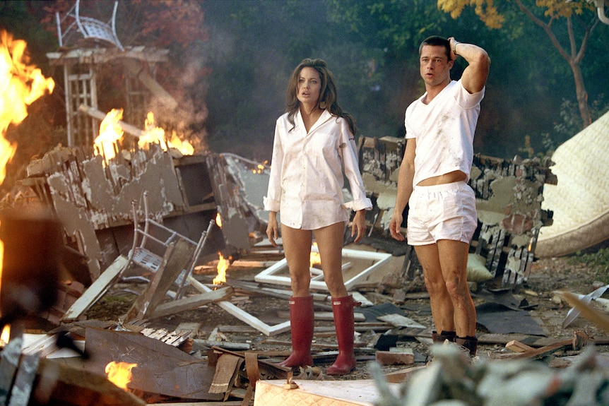 Angelina Jolie wearing a men's shirt with long red boots standing next to Brad Pitt also dressed in white as Mr & Mrs Smith